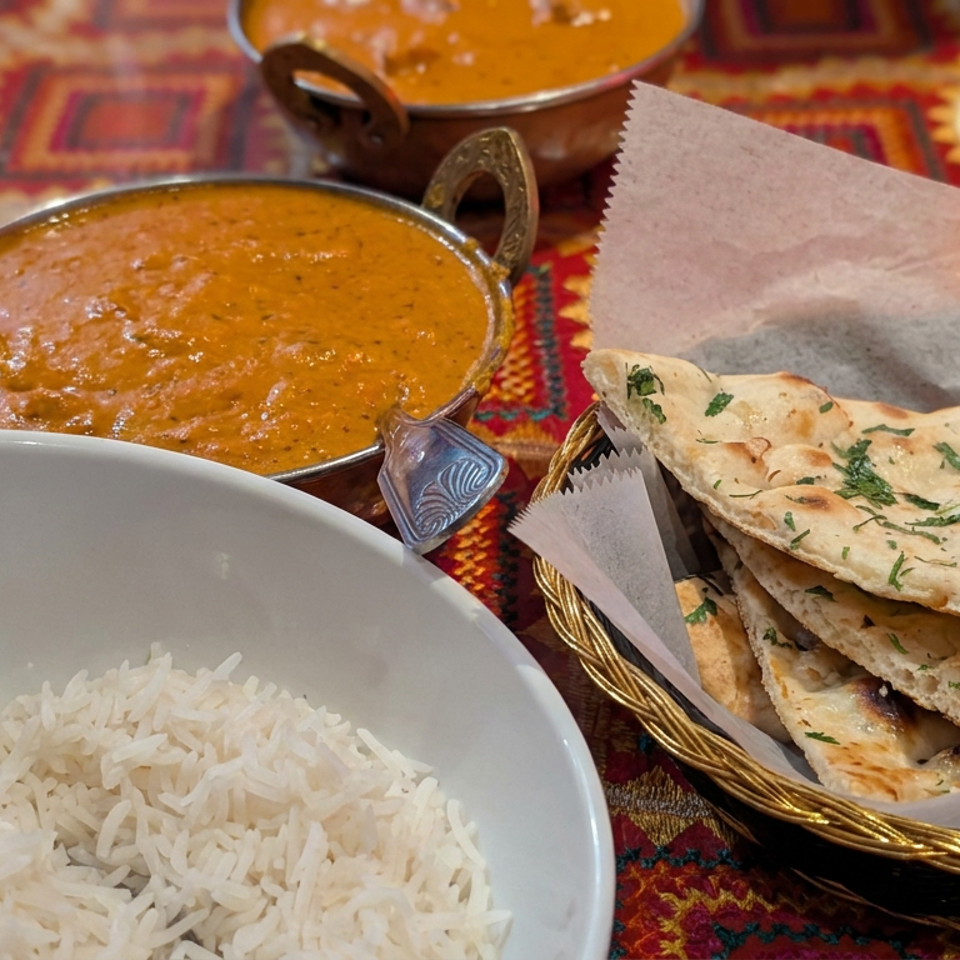 Indian meal featuring basmati rice, two bowls of curry, and a basket of herb-topped naan on a colorful patterned tablecloth.