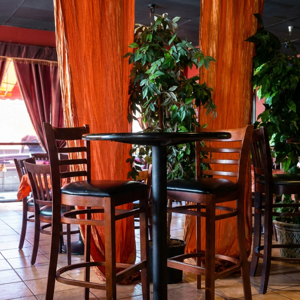 Restaurant dining area with tall wooden tables, barstools, tropical plants, tiled floors, and burgundy curtains.