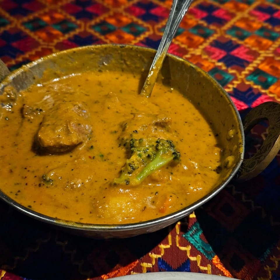 Creamy golden curry with vegetables in a handled metal bowl, served alongside white rice on a colorful patterned tablecloth.